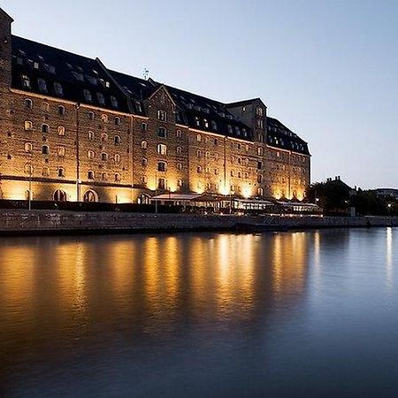 Copenhagen Admiral Hotel Esterno foto The photo shows a large, historic building located beside a calm body of water, likely a river or canal. The structure features a series of tall windows, a dark roof, and is illuminated by warm lights, creating a cozy atmosphere at twilight. Reflecti