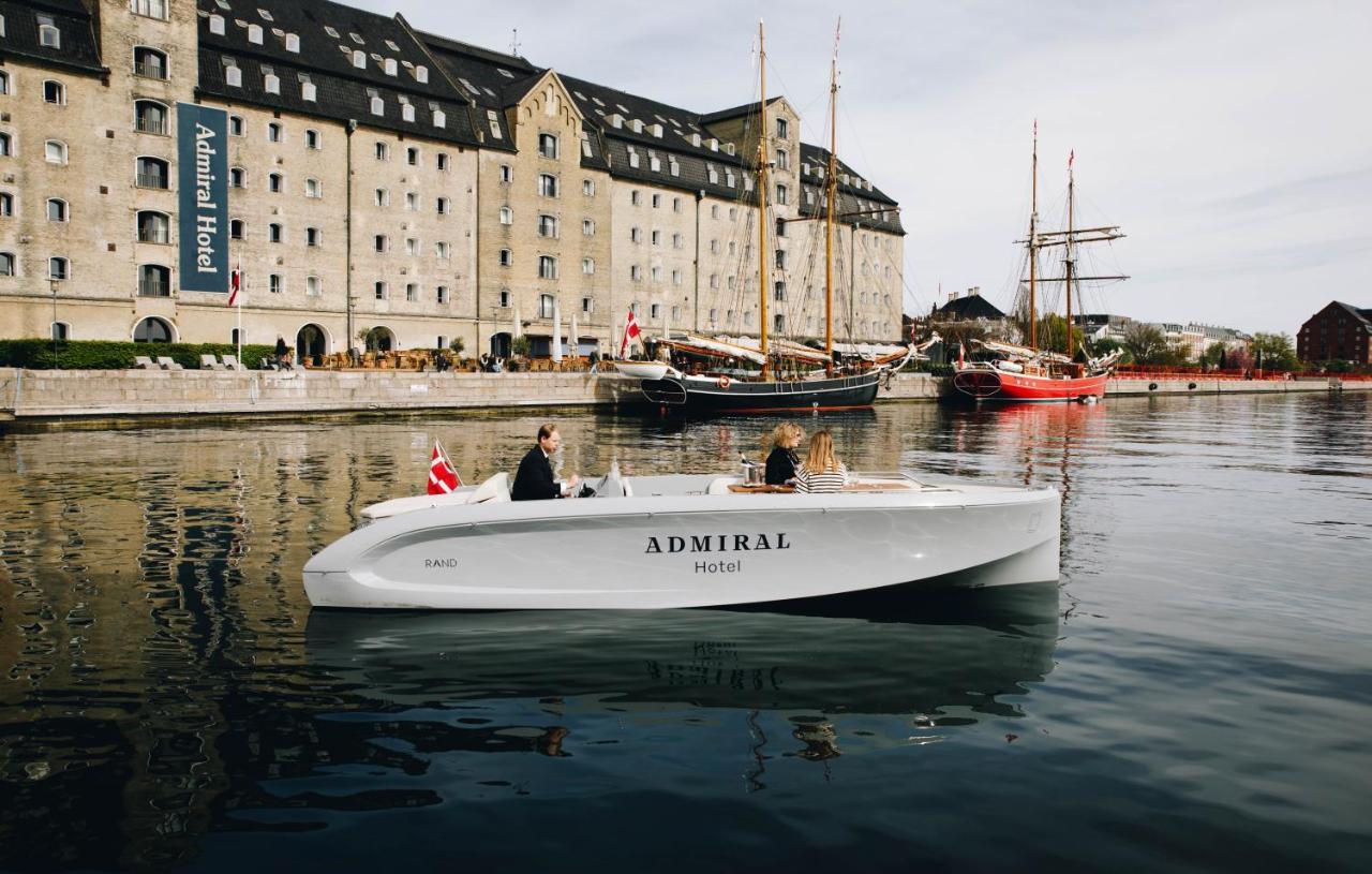 Copenhagen Admiral Hotel Esterno foto The image shows a modern boat with a sleek design labeled "ADMIRAL Hotel" cruising in a calm harbor. In the background, there is a large, historic building that appears to be a hotel, with a distinct architectural style. Several traditional sailboats