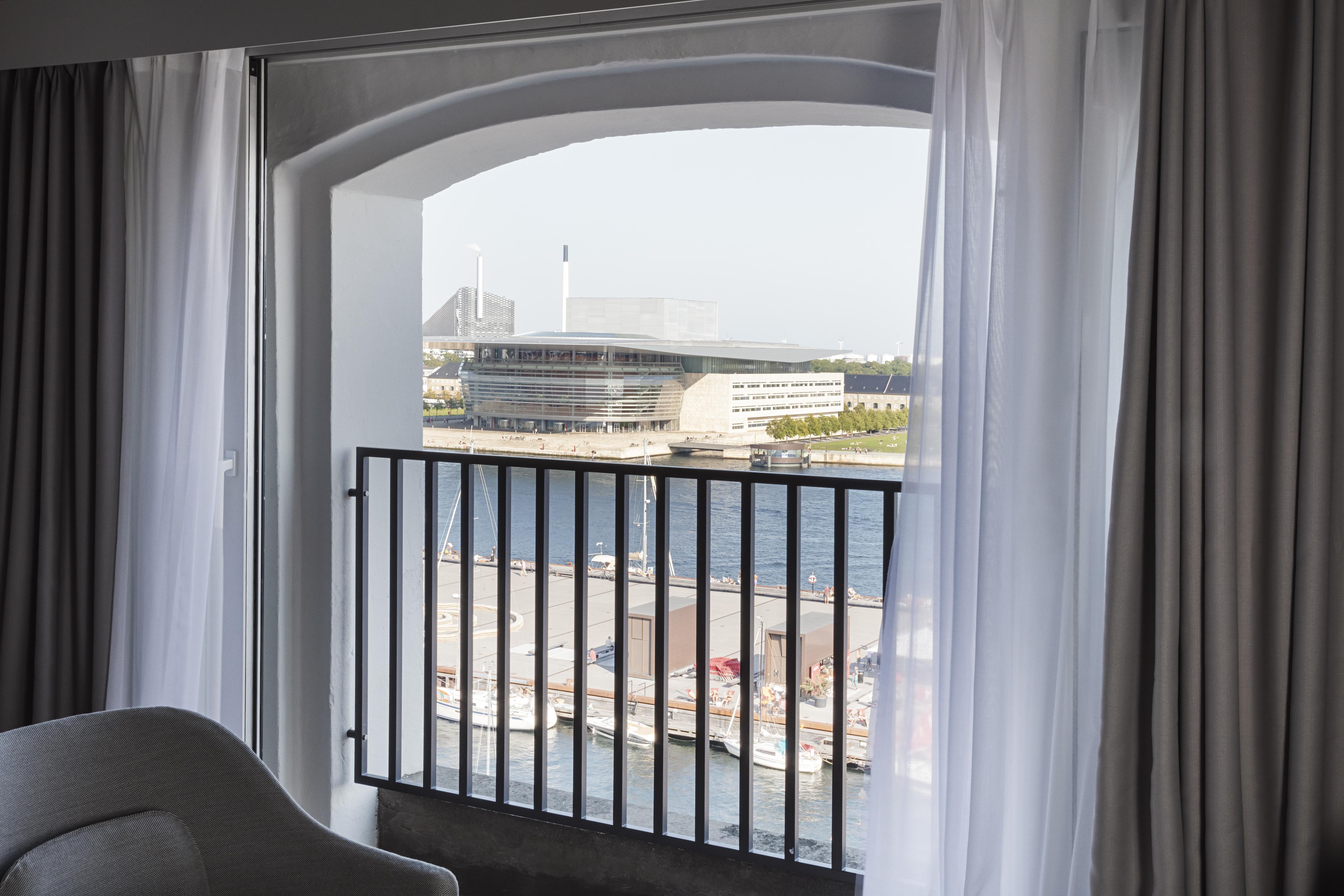 Copenhagen Admiral Hotel Esterno foto The photo shows a view from inside a room looking out through an arched window with sheer curtains. The scene reveals a waterfront area, including a body of water and a modern building in the background, likely a sports or entertainment venue. The ar