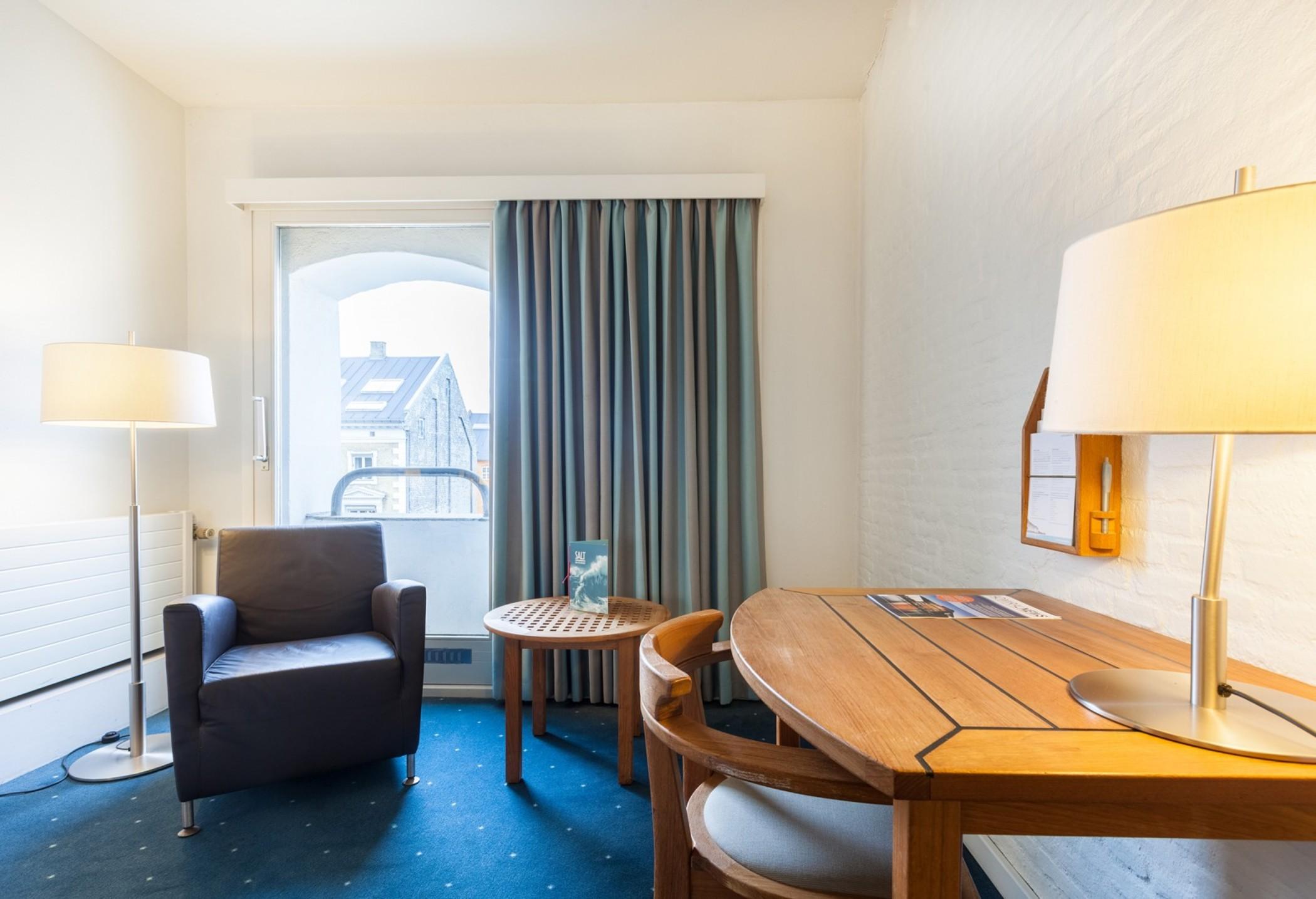 Copenhagen Admiral Hotel Esterno foto The photo shows a small, well-lit hotel room. On one side, there is a blue upholstered armchair next to a round wooden table. The table is accompanied by two wooden chairs. Near the table, there's a lamp providing additional light. A set of curtains 