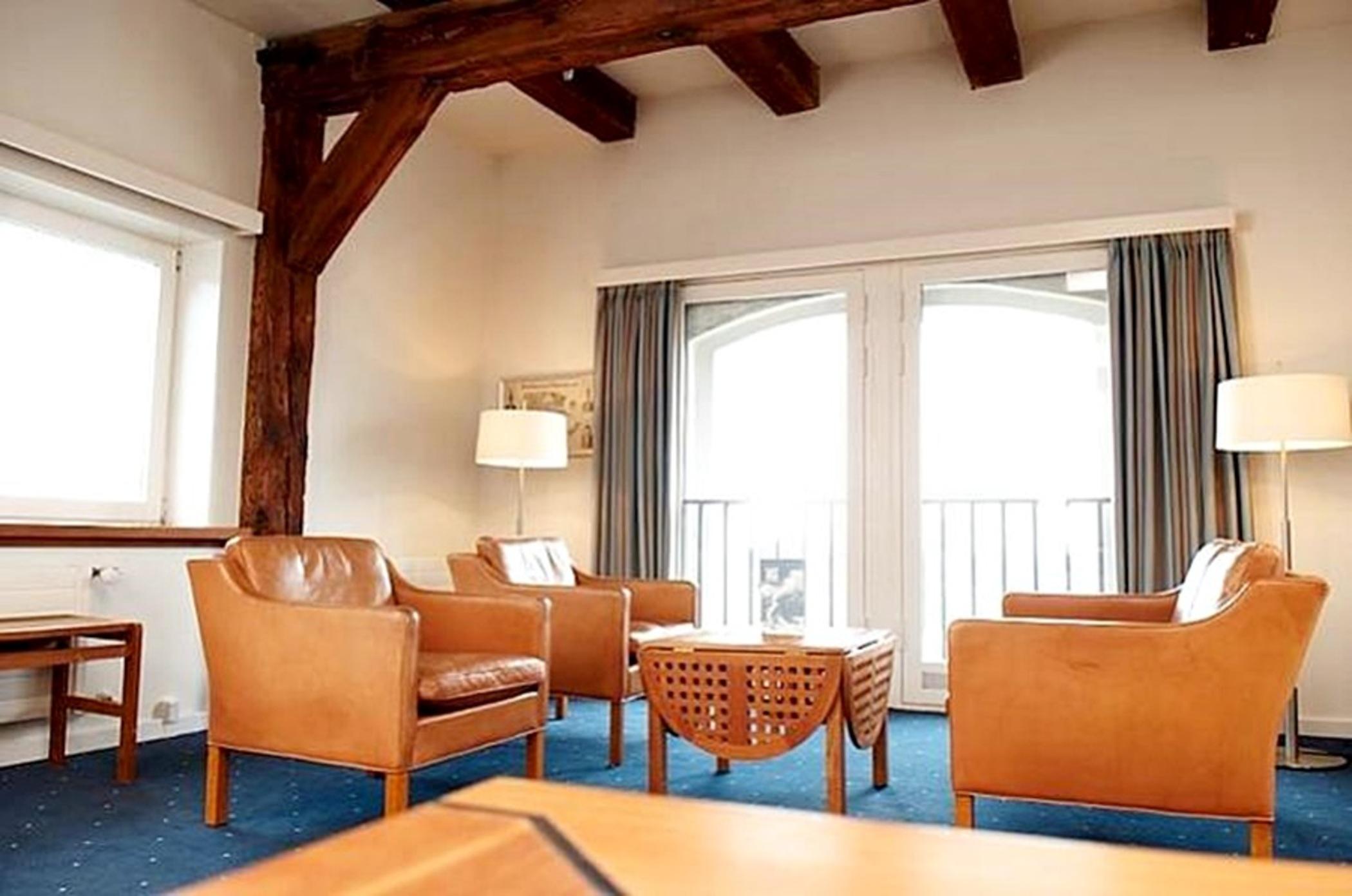 Copenhagen Admiral Hotel Camera foto The photo shows a cozy living area featuring wooden beams on the ceiling. There are four brown leather armchairs arranged around a low, decorative coffee table, which has a geometric design. Two lamps provide lighting, and large windows dressed with 