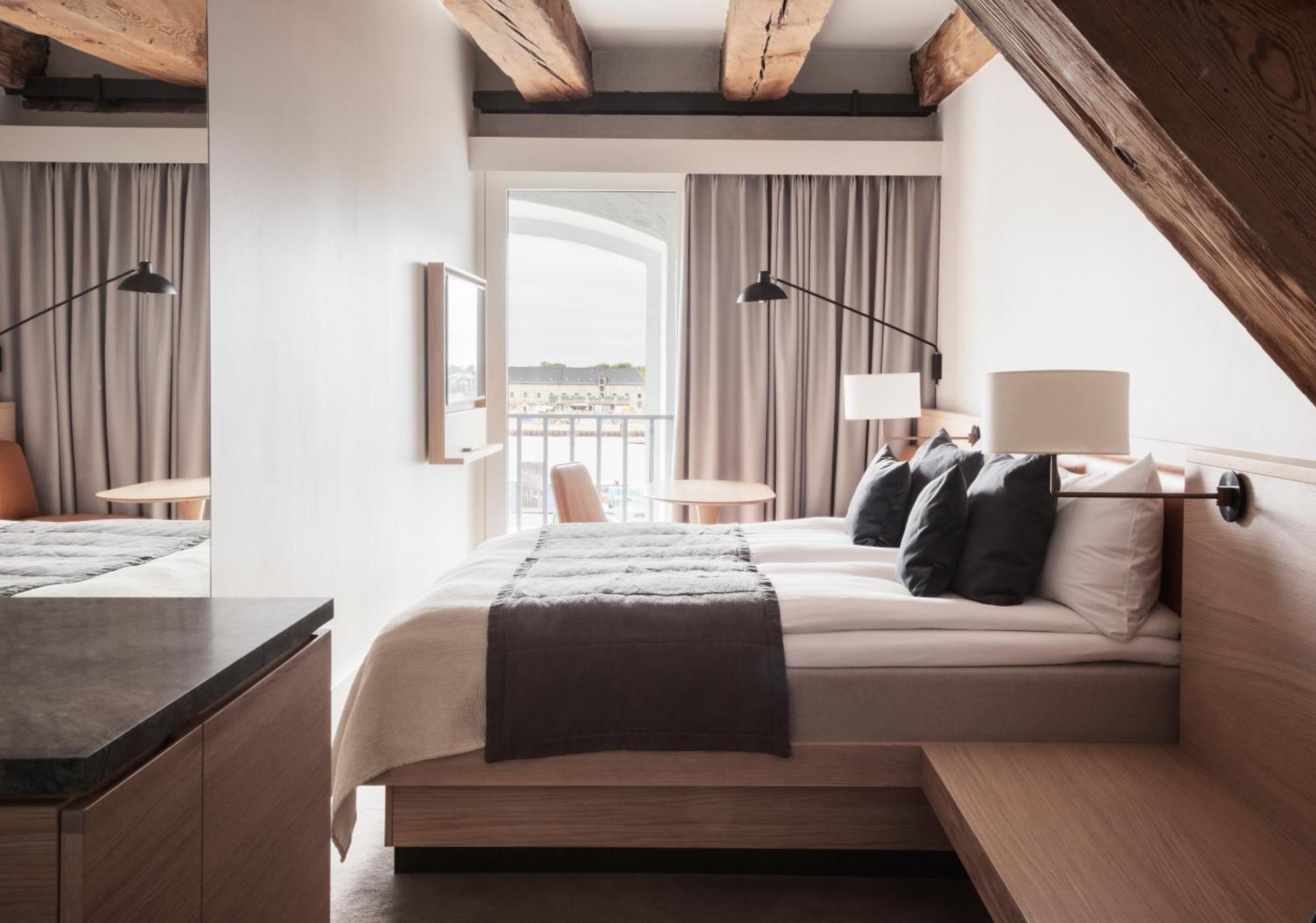 Copenhagen Admiral Hotel Esterno foto The photo shows a modern bedroom with a minimalistic design. It features a large bed with white linens and black accent pillows. There is a bedside table on one side and a small sitting area with a table and chairs visible through a large window. The
