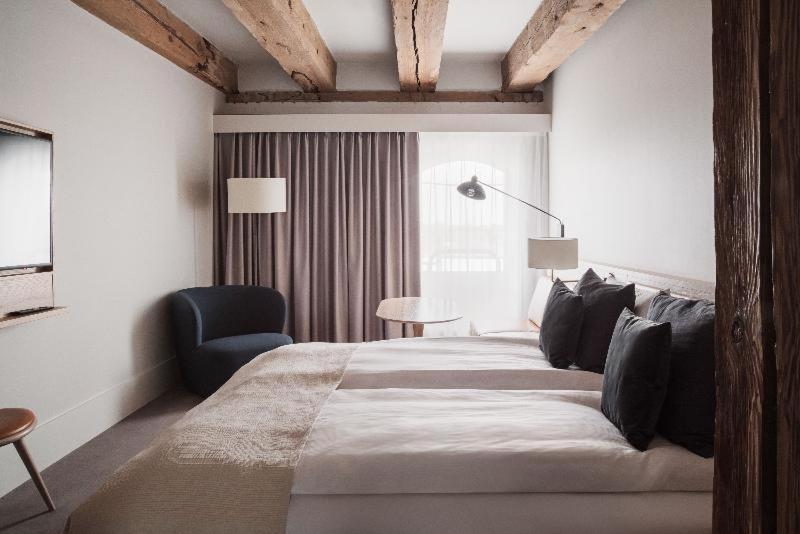 Copenhagen Admiral Hotel Camera foto The photo depicts a modern hotel room featuring two beds with white bedding and dark pillows. There is a cozy chair in the corner, alongside a small round table. Large curtains in a neutral tone cover the window, and a stylish floor lamp provides add