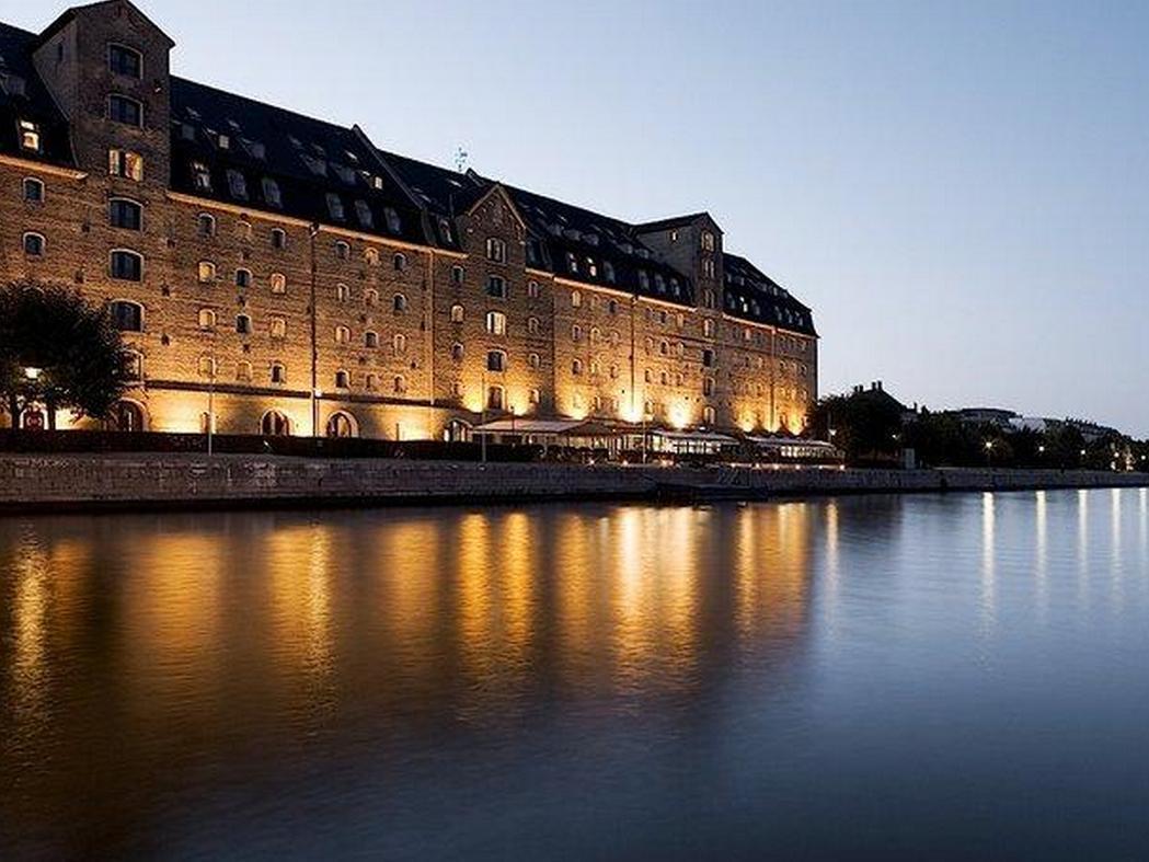 Copenhagen Admiral Hotel Esterno foto The photo shows a large, historic building located beside a calm body of water, likely a river or canal. The structure features a series of tall windows, a dark roof, and is illuminated by warm lights, creating a cozy atmosphere at twilight. Reflecti