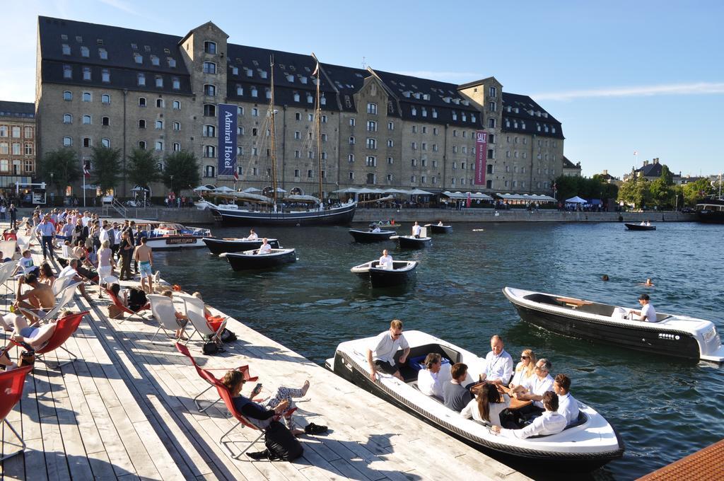 Copenhagen Admiral Hotel Esterno foto The photo depicts a lively waterfront scene with several boats in the water. People are enjoying their time on the shore, some lounging in deck chairs while others are seated in boats. In the background, a large building with a distinctive architectu