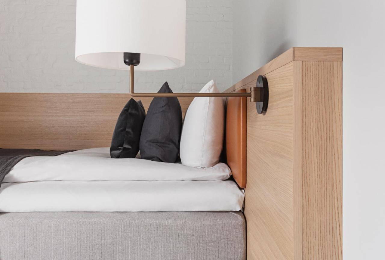 Copenhagen Admiral Hotel Esterno foto The photo shows a modern bedroom setting. It features a bed with a wooden headboard, adorned with a few neatly arranged pillows. There are two black pillows and one white pillow on the bed. A sleek, stylish lamp is mounted on the side of the headboar