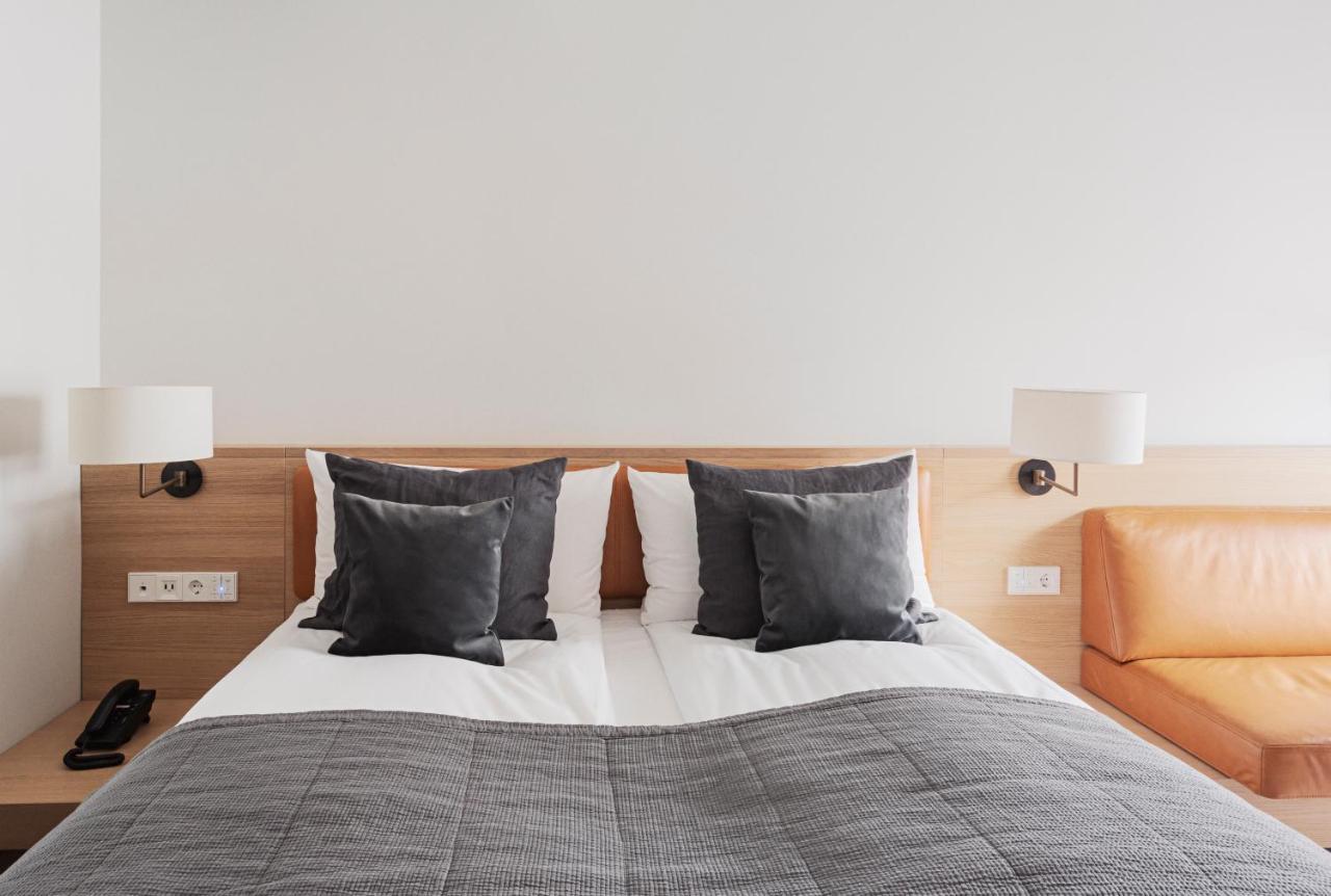 Copenhagen Admiral Hotel Esterno foto The photo shows a modern hotel room featuring a neatly made bed with white linens and two dark gray decorative pillows. The bed is positioned against a light-colored wooden panel wall. On either side of the bed, there are wall-mounted lamps providing