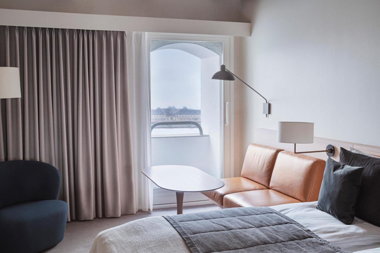 Copenhagen Admiral Hotel Esterno foto The photo shows a modern hotel room with a clean and minimalist design. There is a bed with a neatly arranged gray blanket and several pillows. Beside the bed is a small table with a lamp. A light brown leather sofa is positioned near a window, which