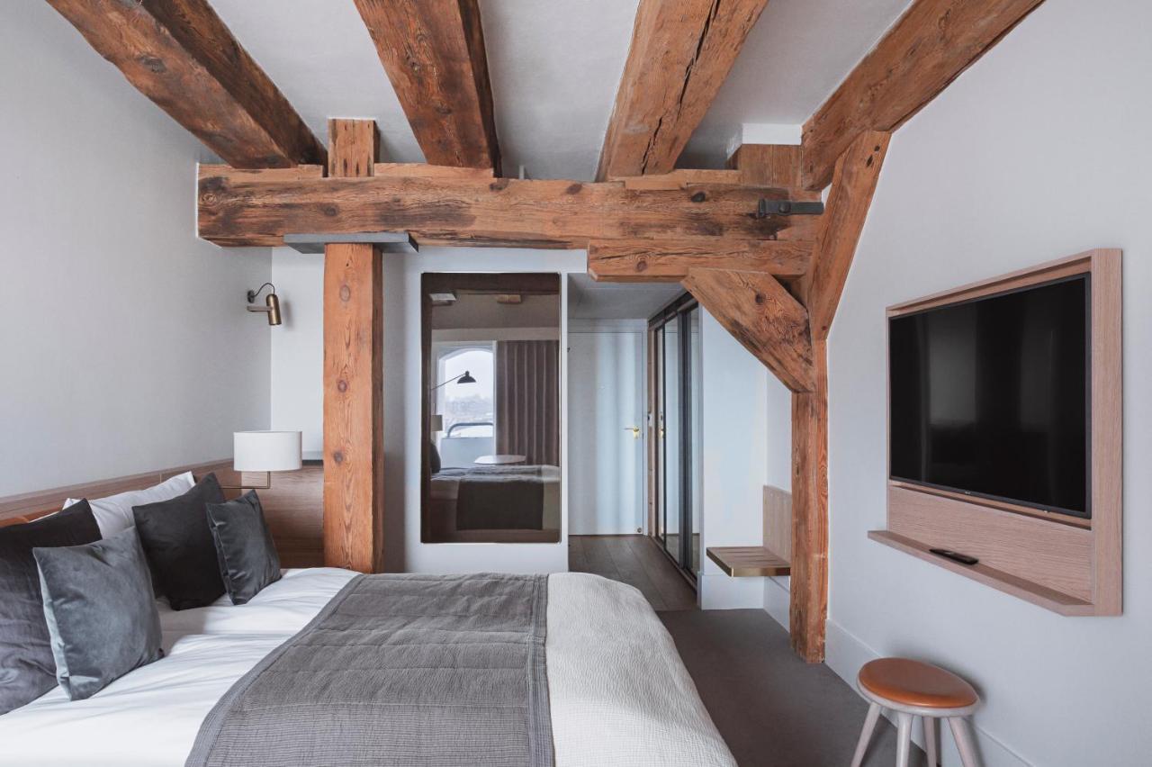 Copenhagen Admiral Hotel Esterno foto The photo depicts a modern bedroom featuring a cozy design. There is a large bed with pillows and a gray throw blanket. Exposed wooden beams are visible on the ceiling, adding a rustic touch to the room. A minimalist nightstand with a lamp is next to